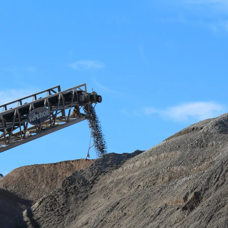 Industrial conveyor belt dumping gravel onto a large pile under a clear blue sky outdoors