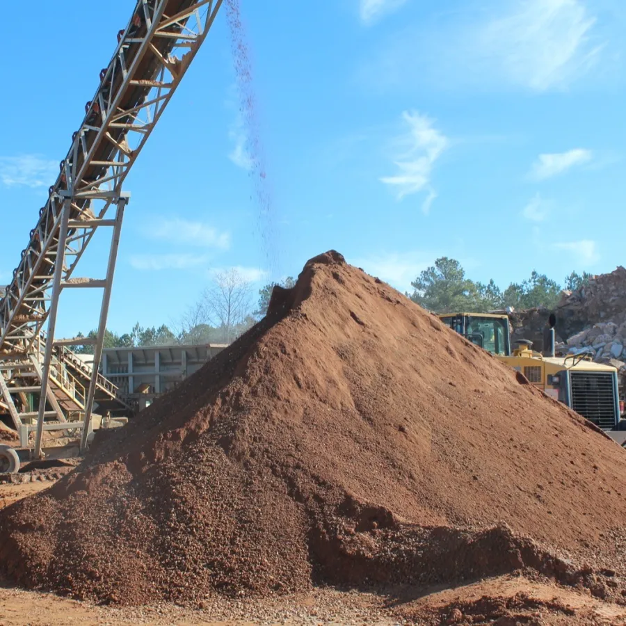 Conveyor belt dumping reddish gravel into a large pile at an outdoor mining or construction site under clear sky.
