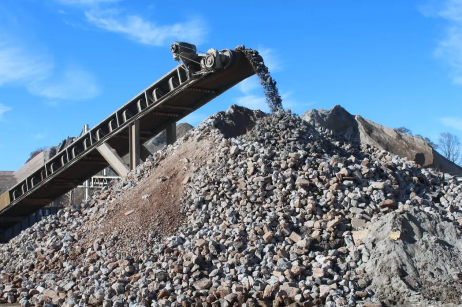 Conveyor belt dumping crushed stones on a large rocky pile under a clear blue sky at a quarry site.