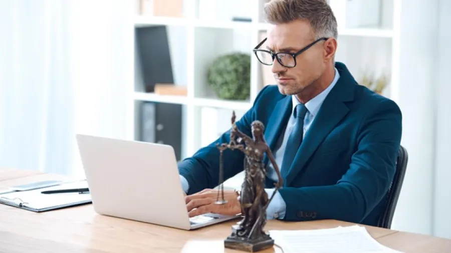 Professional man in glasses working on laptop at desk with Lady Justice statue and legal documents nearby
