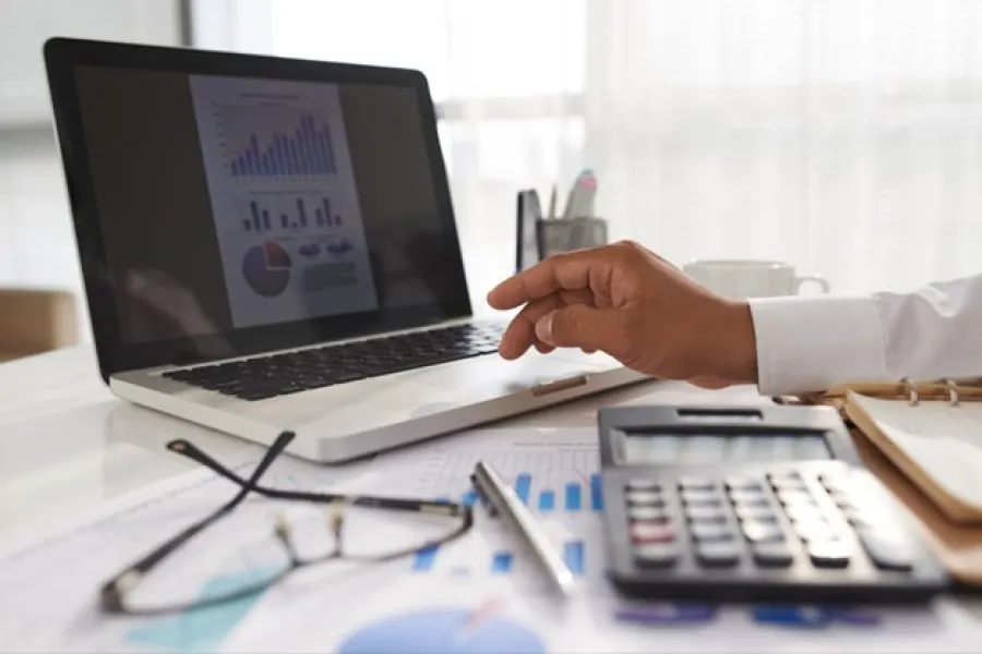 Person analyzing financial charts on laptop with calculator, pen, and glasses on desk in office setting