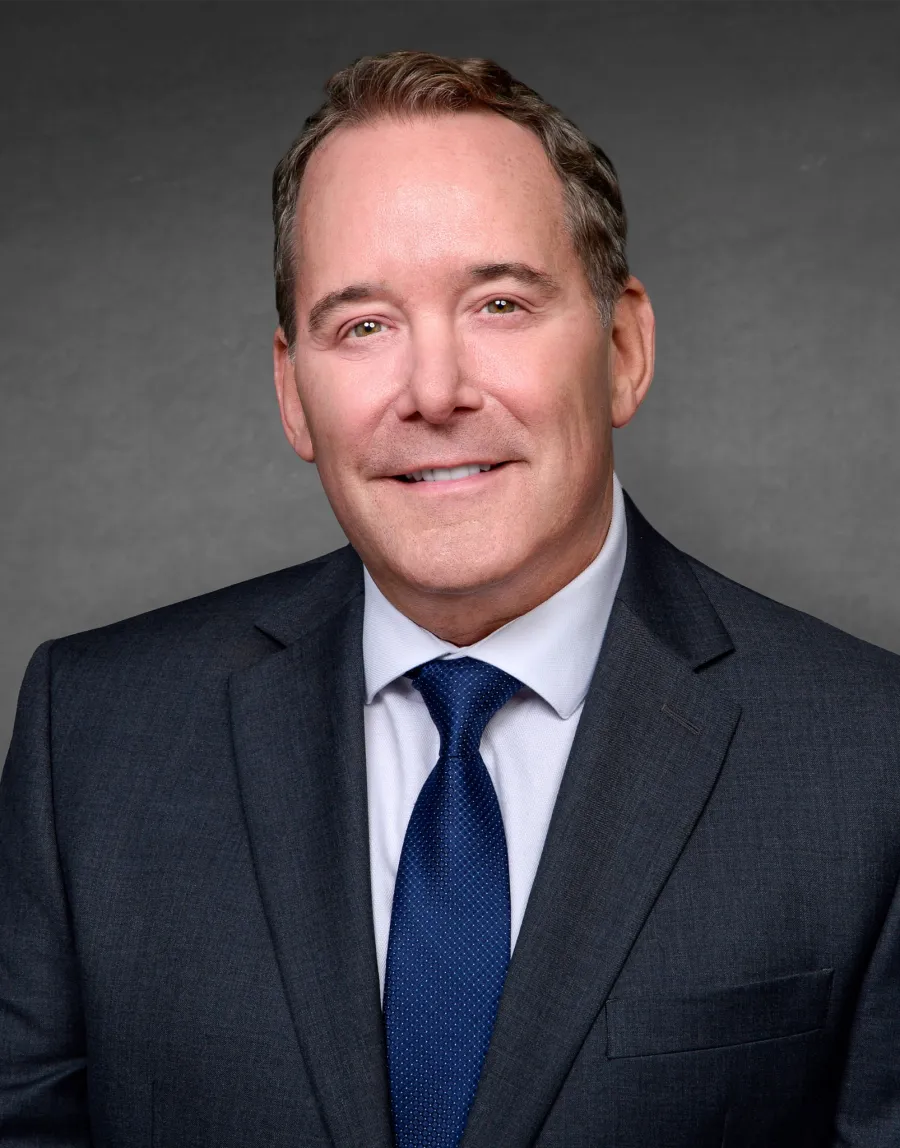Smiling middle-aged man in dark suit jacket, white shirt, and blue tie against gray backdrop.