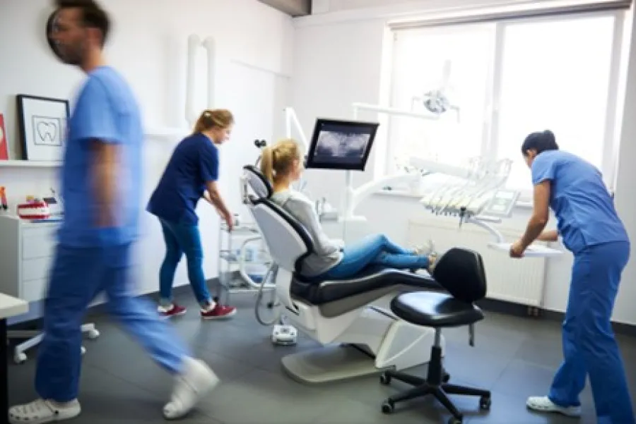 Dental clinic with patient in chair and three staff members in blue scrubs attending and preparing equipment.