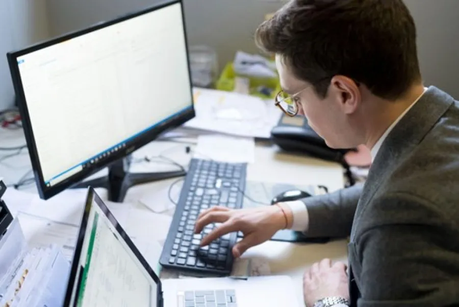 Man in glasses working at a desk using a laptop and desktop with spreadsheets and documents scattered around.