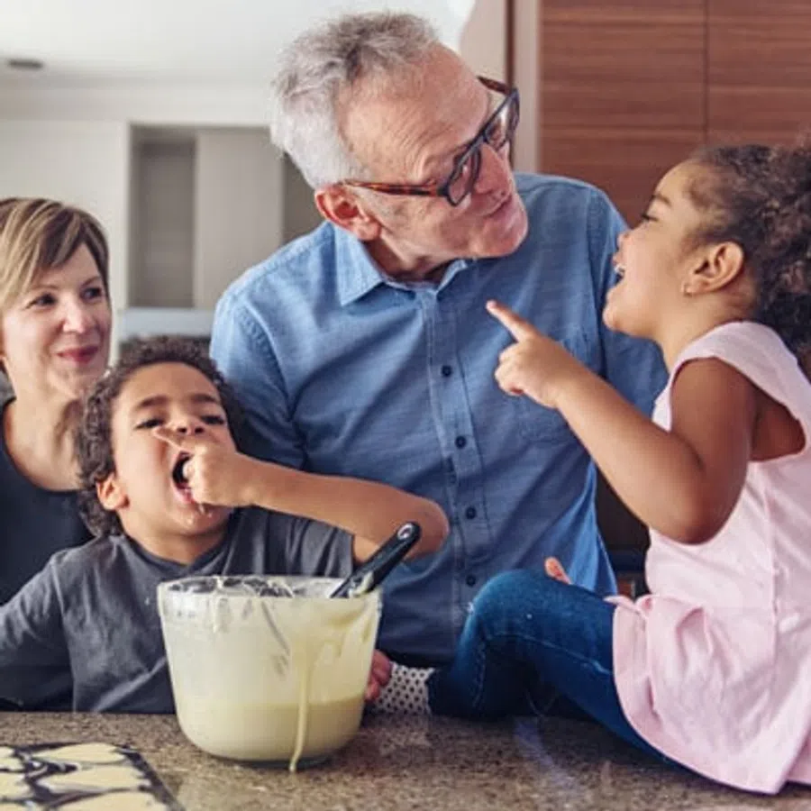 Family baking together, children tasting batter and laughing with grandparents in kitchen setting.
