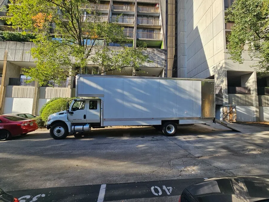 White delivery truck with open ramp parked on city street beside a red car during daytime.