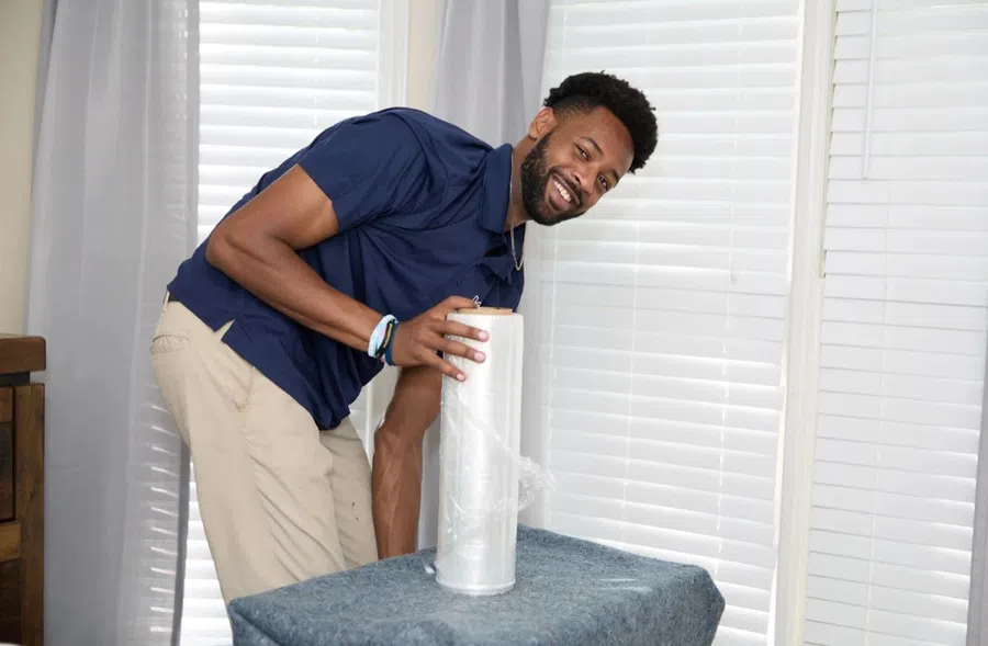 Smiling man preparing furniture with plastic wrap in a bright room with closed white blinds.