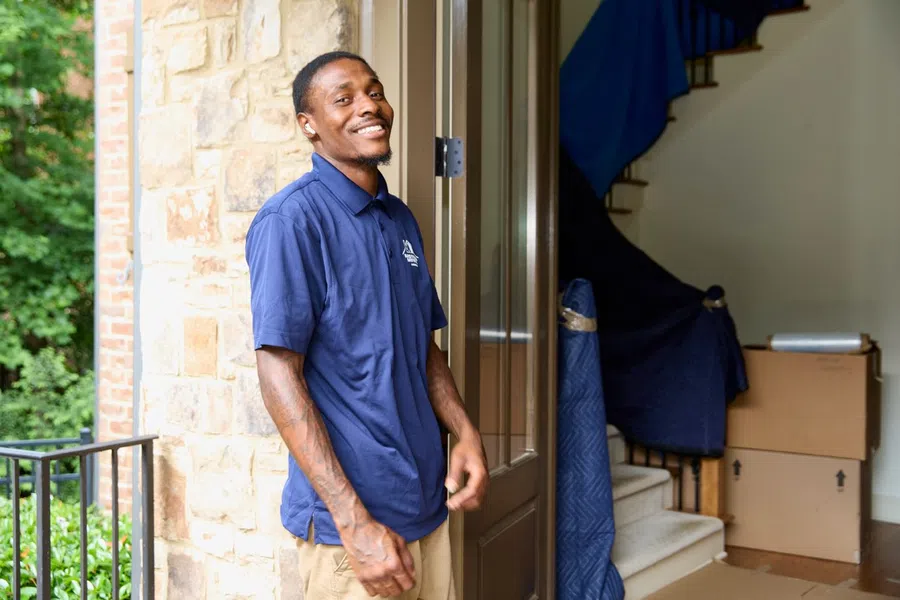 Smiling mover in blue shirt standing by an open door with packed boxes and covered furniture inside.