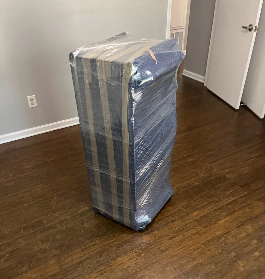 Tall rectangular furniture wrapped in protective plastic wrap and moving blankets on wooden floor indoors.