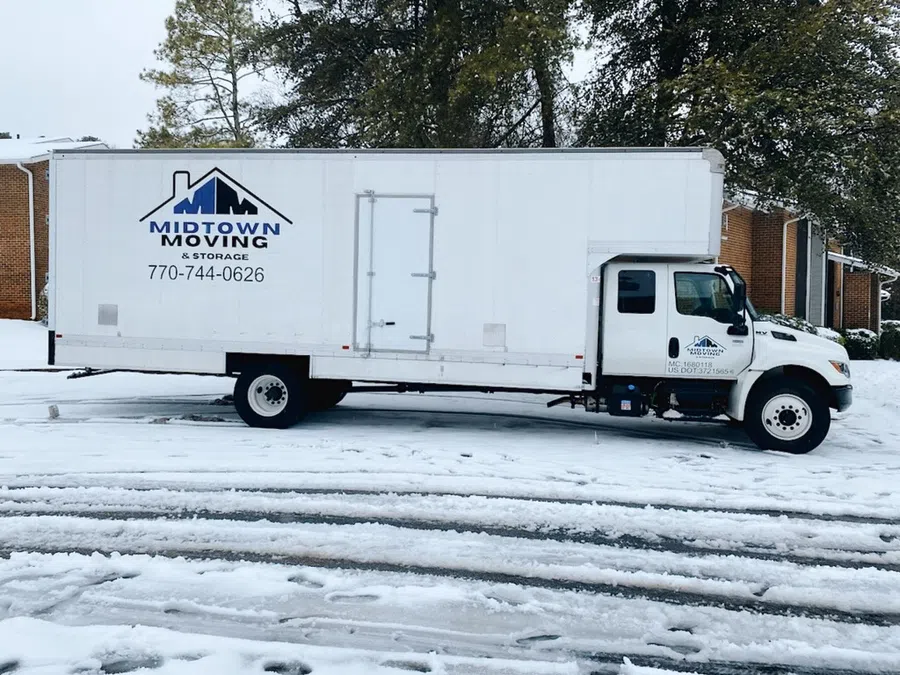 White Midtown Moving truck parked on snowy ground near trees and brick buildings on a cloudy day