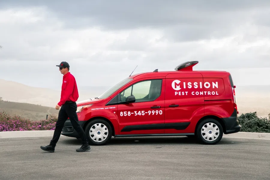Red Mission Pest Control van parked on road with technician in red uniform walking nearby under cloudy sky.