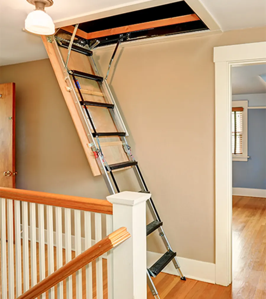 Retractable attic ladder extended from ceiling into a bright home hallway with wooden flooring.