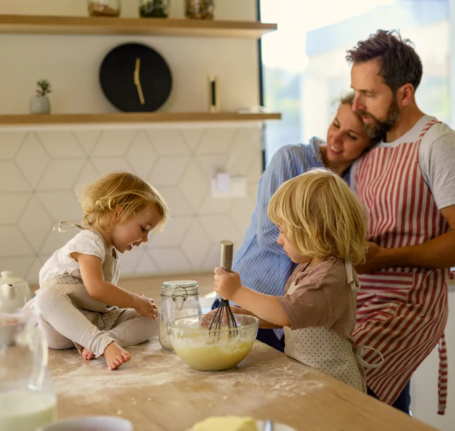 Parents watch two young children mixing batter in a kitchen with a wooden countertop and modern decor.