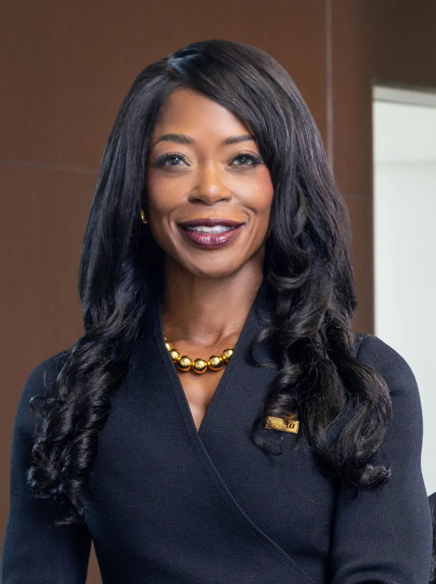 Professional woman with long curly hair wearing a black outfit and gold necklace smiling indoors.