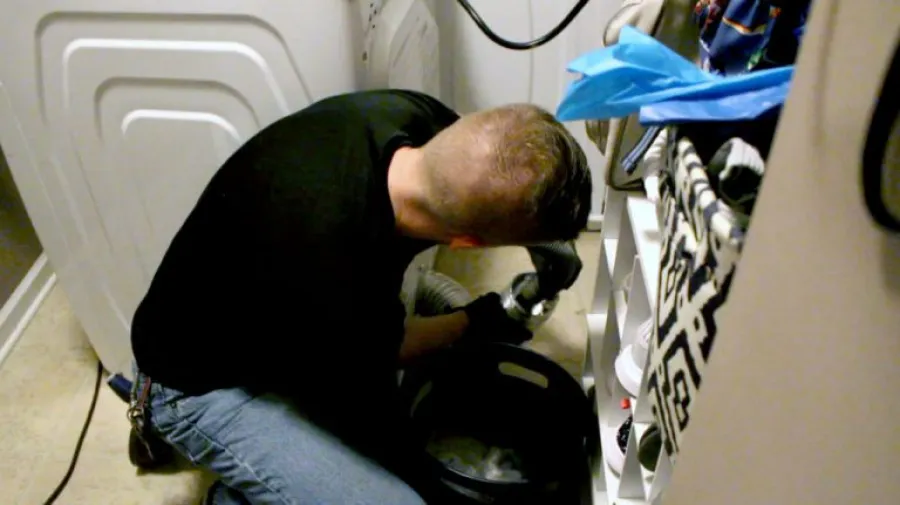 Man wearing black shirt and gloves installs or repairs a washing machine in a laundry room next to a shoe rack.