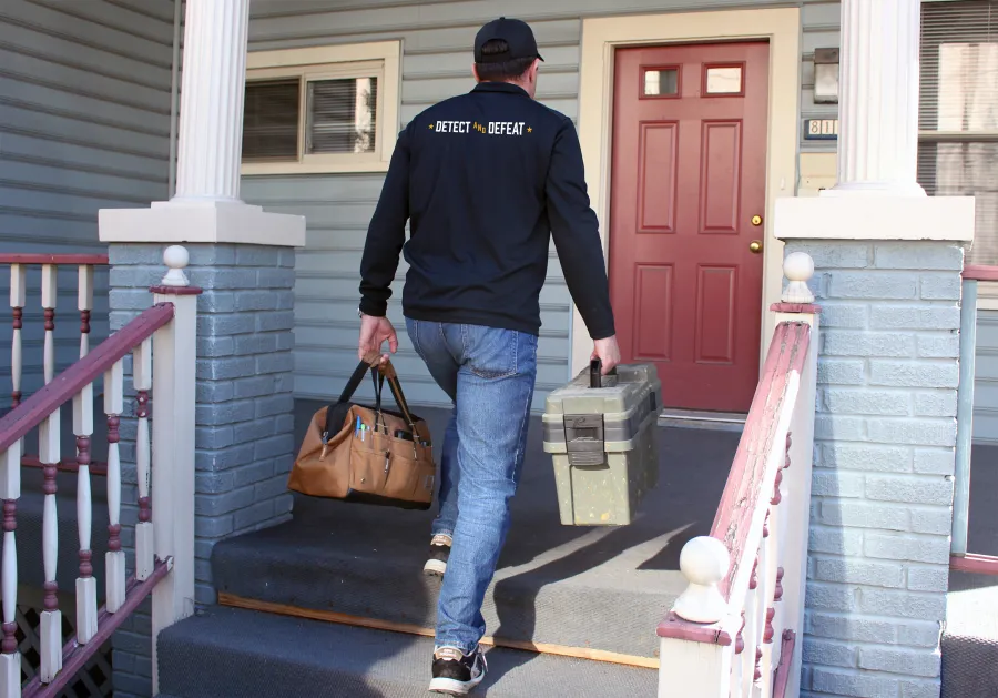 Technician wearing a Detect and Defeat jacket carrying toolboxes up the porch stairs to a red front door.