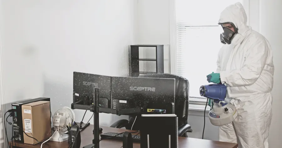 Person in protective suit disinfecting a home office with multiple monitors and a window in daylight.