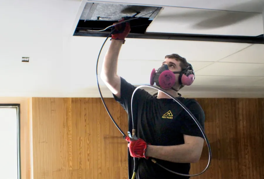 Technician wearing a respirator installs or inspects ceiling ductwork in an indoor room with wooden panels.