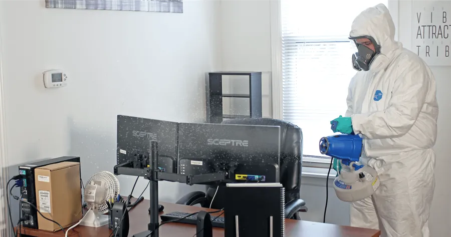 Worker in protective suit and respirator cleaning wooden shelves with industrial vacuum hose indoors.