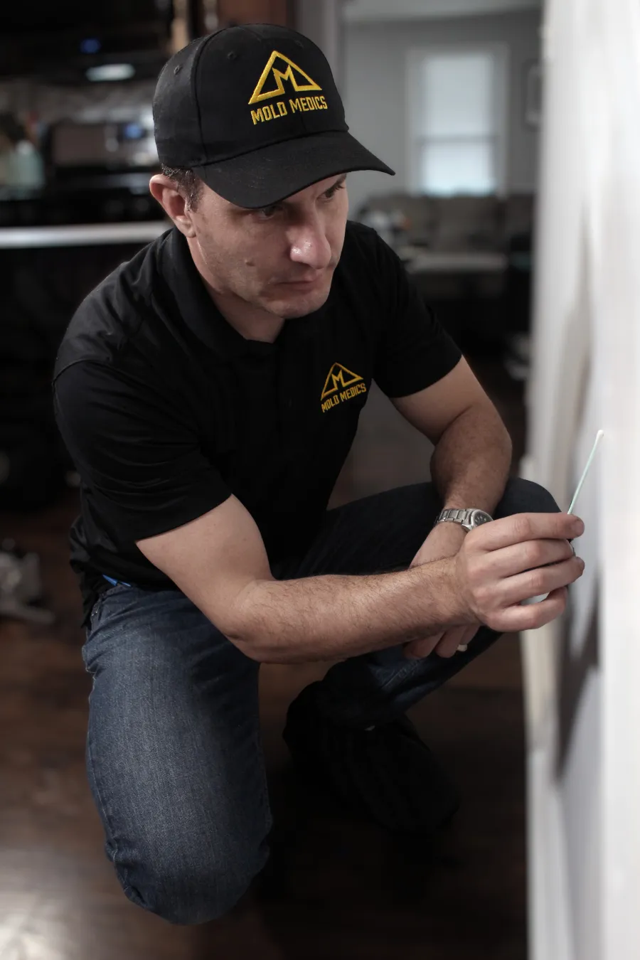 Technician in black Mold Medics uniform inspecting a wall with a testing swab inside a home.