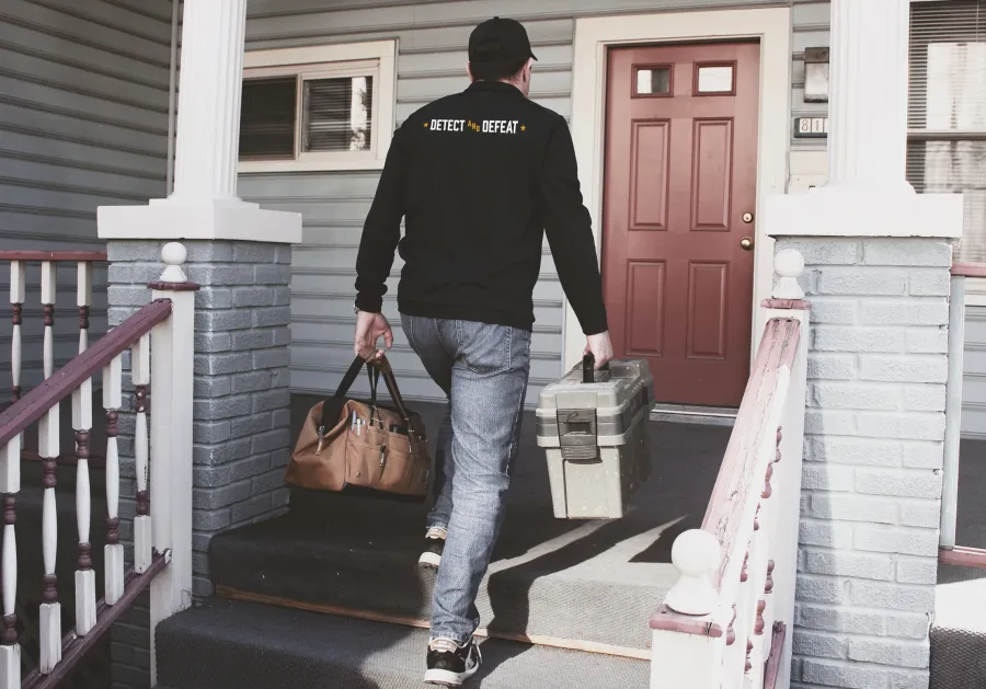 Technician wearing Detect and Defeat jacket carrying tool bags up porch steps to a red front door of a house.