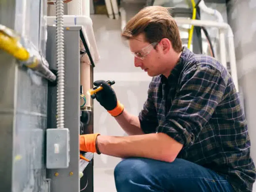 Technician in safety gloves and glasses inspecting HVAC system with flashlight in a utility room