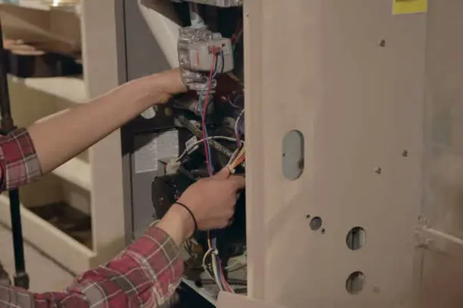 Technician repairing HVAC system with tools and wiring inside a furnace unit in a utility room.