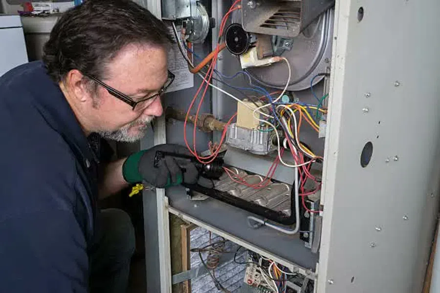Technician inspecting and repairing a home furnace with tools and exposed wiring inside the unit.
