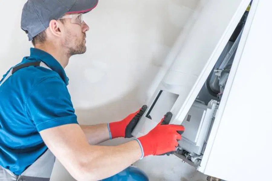 Technician in red gloves and cap installing or repairing a white wall-mounted boiler or heater indoors.