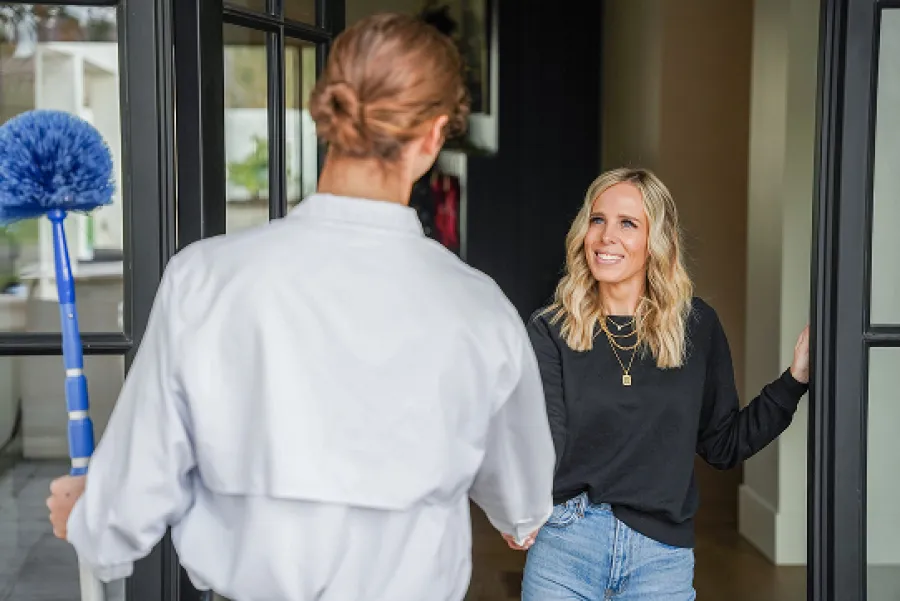 Smiling woman greets a cleaning professional holding a blue duster at her home entrance.