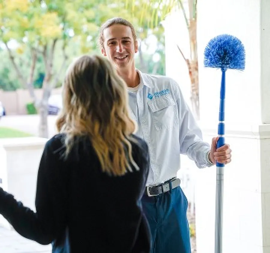Technician in uniform holding a long blue brush speaks to a woman in casual clothes outdoors near a white building.