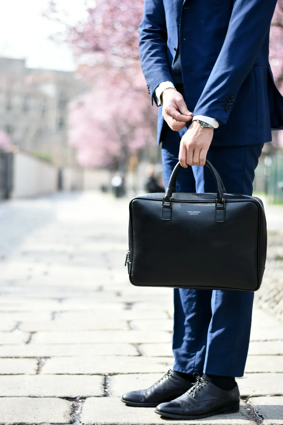 Businessman in blue suit holding black leather briefcase standing on cobblestone street with cherry blossoms.
