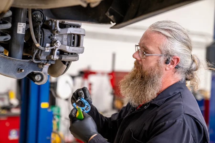 Bearded mechanic wearing glasses and gloves inspecting car brake components in an auto repair shop.
