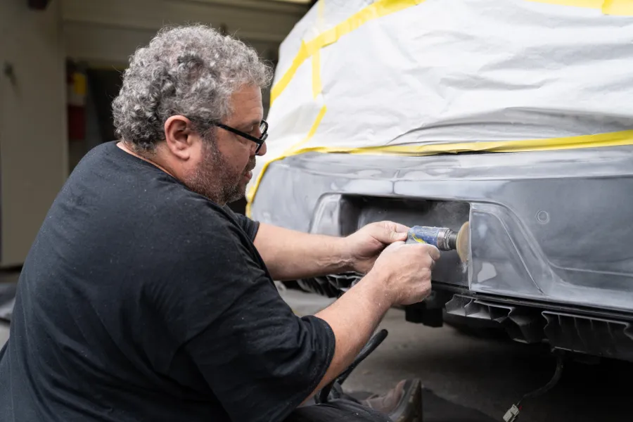 Man sanding a car bumper in a workshop with protective masking tape applied on the car body.