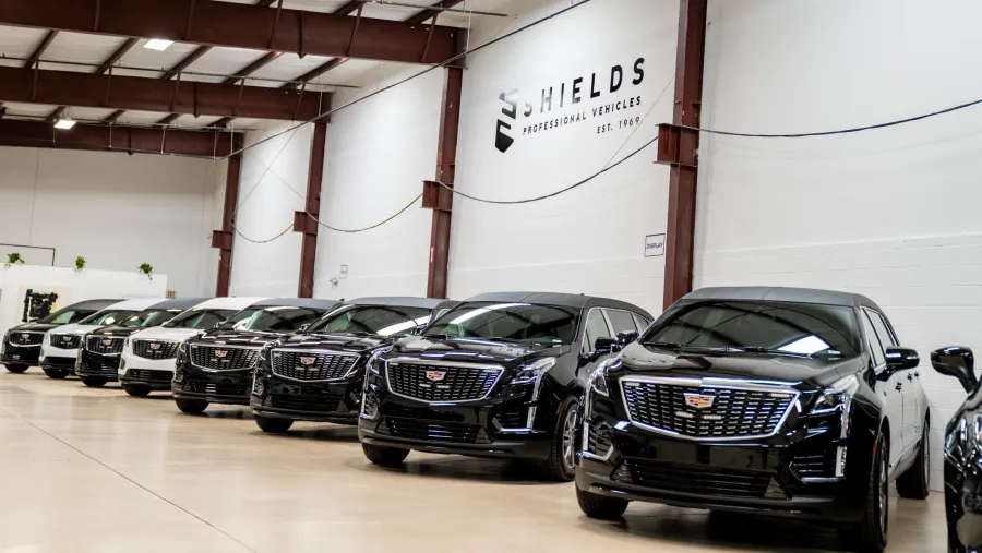 Row of black and white Cadillac SUVs inside a large warehouse with Shields Professional Vehicles branding.