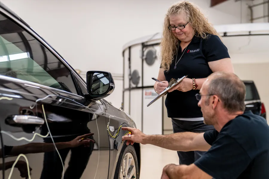 Man inspecting a car door with a tool while a woman takes notes on a clipboard in a garage.