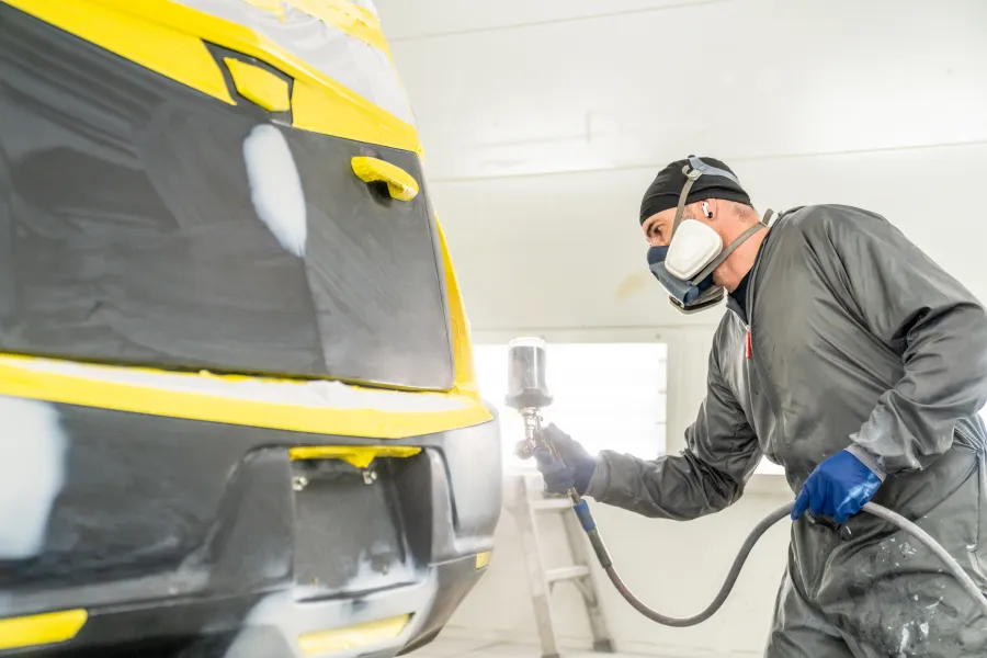 Auto body technician wearing protective gear spray painting a car's rear section in a workshop.
