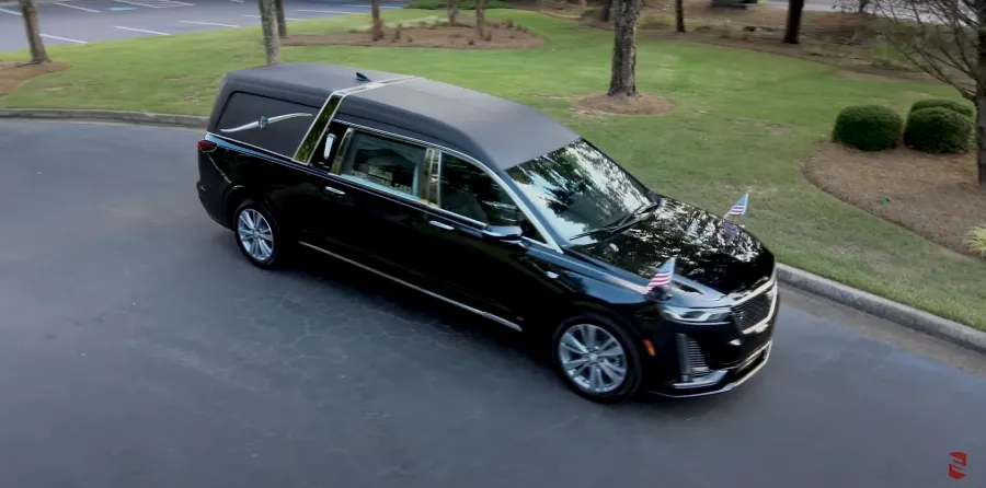 Black hearse with U.S. flags parked on a smooth surface surrounded by trees.