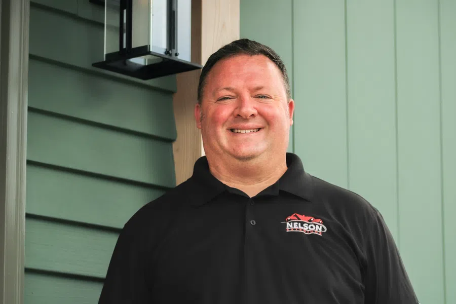 Smiling man wearing a black polo with Nelson logo standing against green siding and wooden trim outdoors.