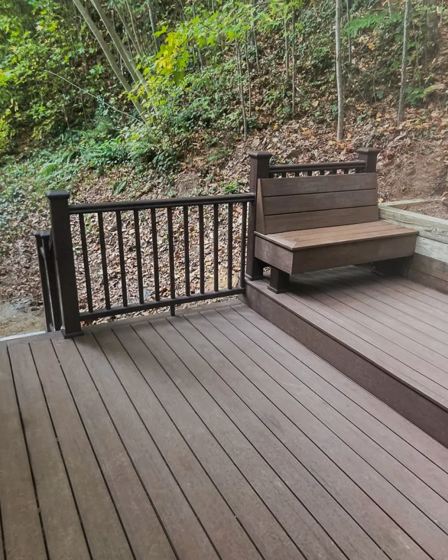 Outdoor wooden deck with built-in bench and railing surrounded by green and leafy hillside.