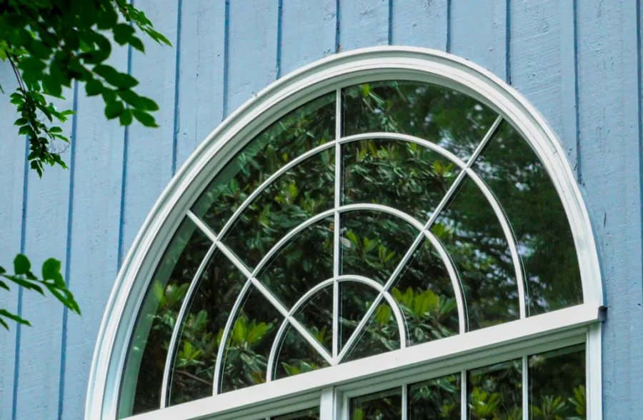 Arched white-framed window reflecting green leafy trees on blue wooden siding house exterior.