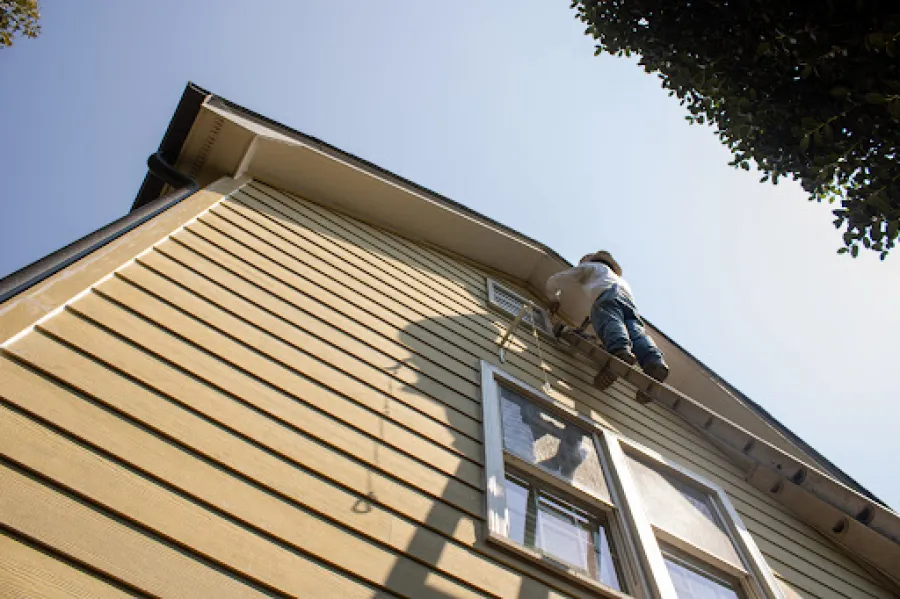 Two-story white house with green roof, front porch, stairs, surrounded by bushes and a clear blue sky.