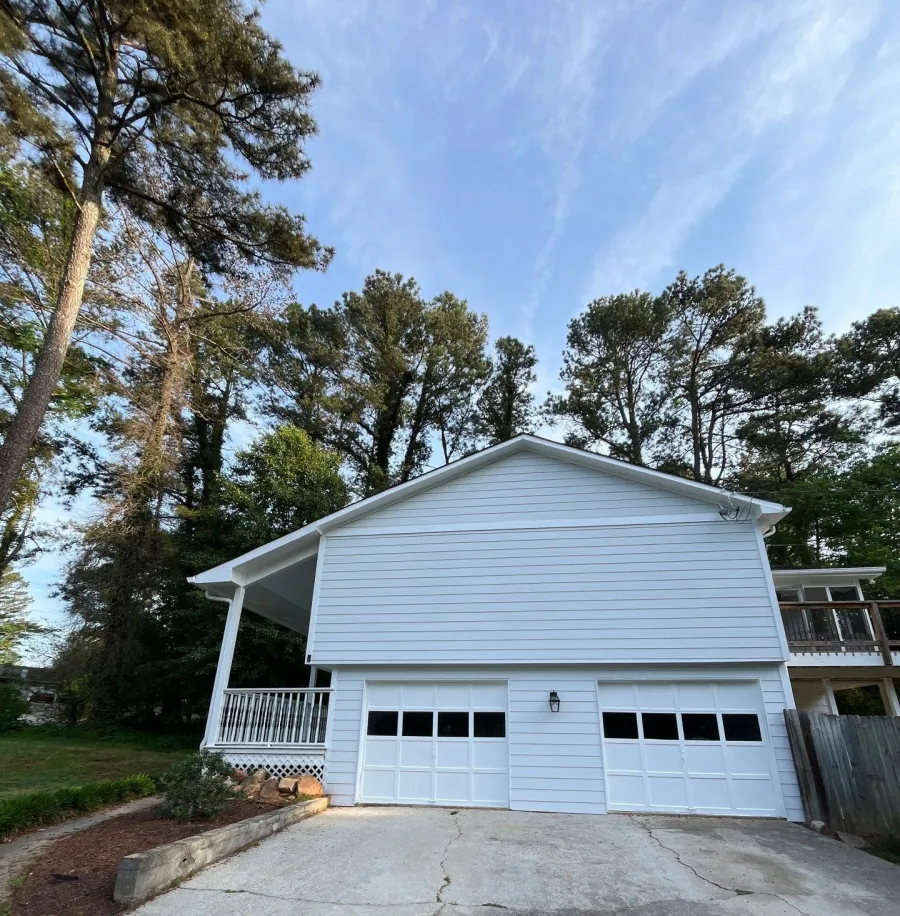 White two-car garage attached to a house with a concrete driveway and surrounded by tall trees under a blue sky