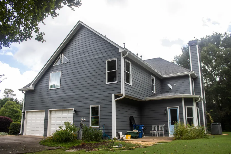 Two-story gray house with white trim, double garage, backyard patio, and surrounding greenery under a cloudy sky