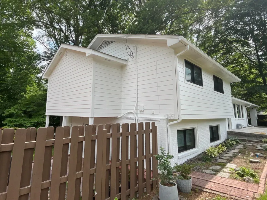 Two-story white house with black windows, surrounded by trees and a brown wooden fence in the front yard.