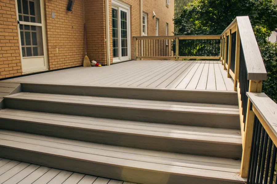 Wide view of a clean wooden deck with stairs and railing attached to a brick house under sunlight.