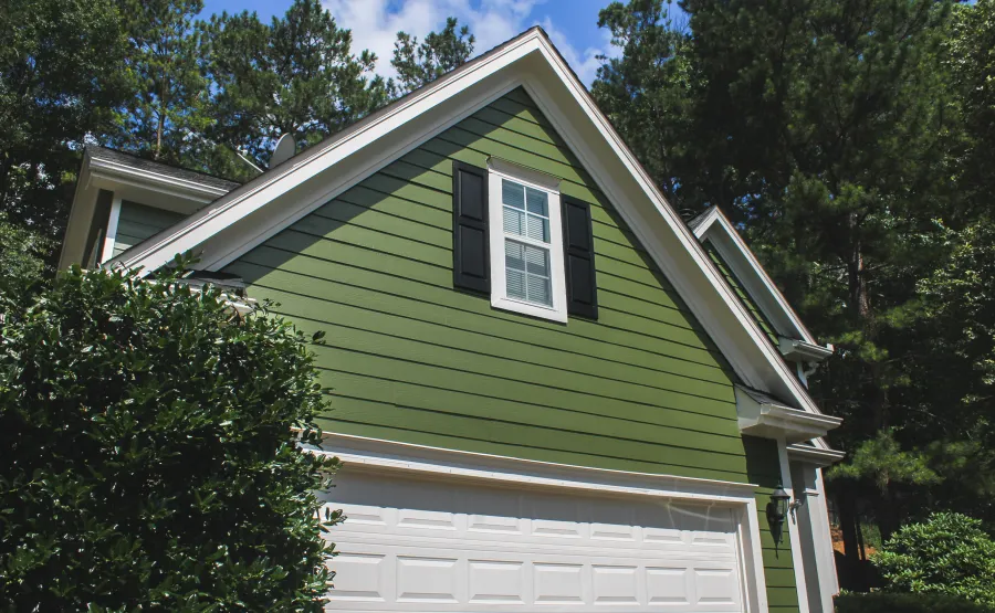 Green house exterior with white garage door, black window shutters, surrounded by trees and blue sky.