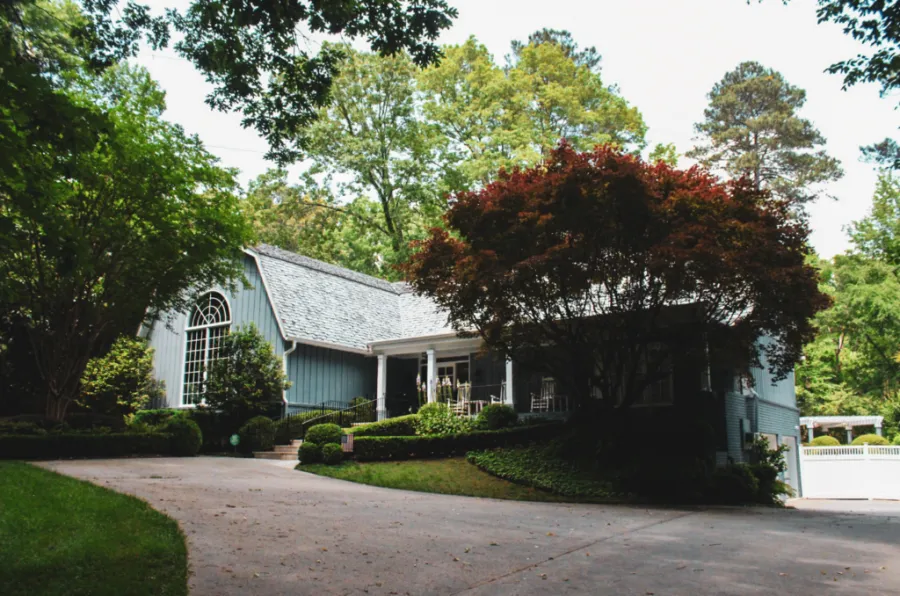 Cozy blue house surrounded by lush trees and greenery with a curved driveway and front porch seating.