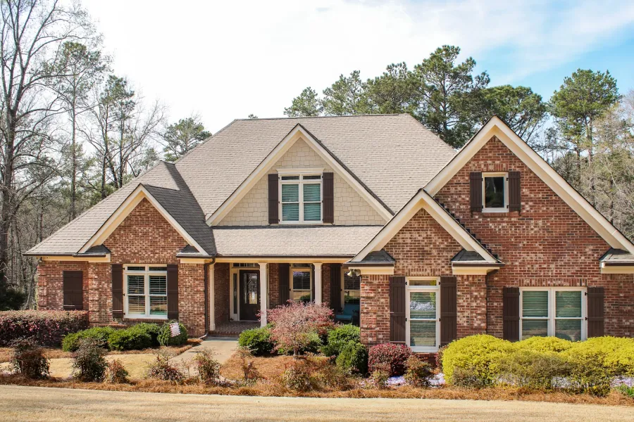 Large brick suburban house with gabled roof surrounded by shrubs and leafless trees under blue sky.