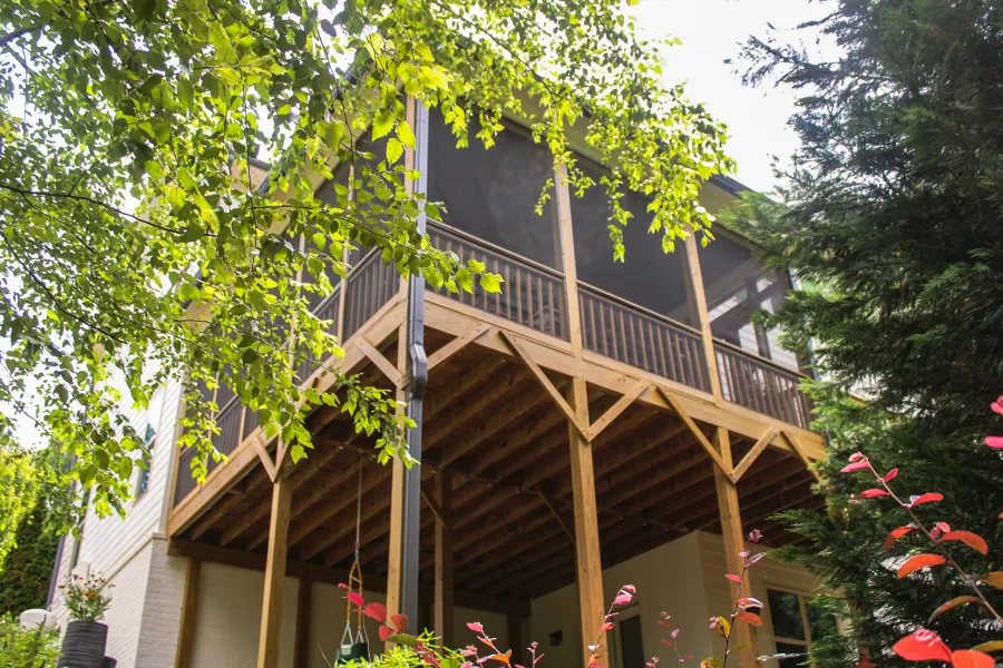 Elevated wooden deck with screened porch surrounded by lush green trees and colorful plants.